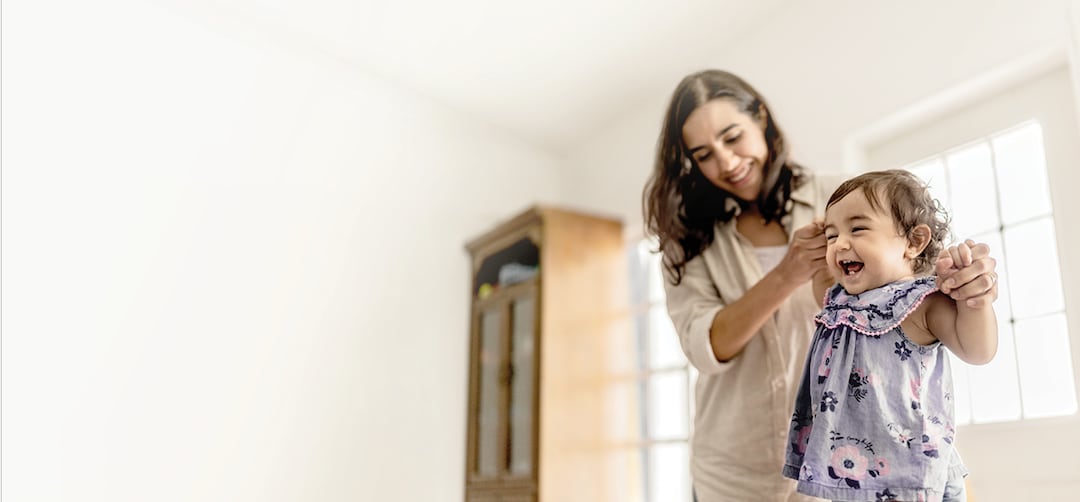 An image of a smiling young woman, possibly a mother, holding up and trying to steady a standing toddler who has a big smile